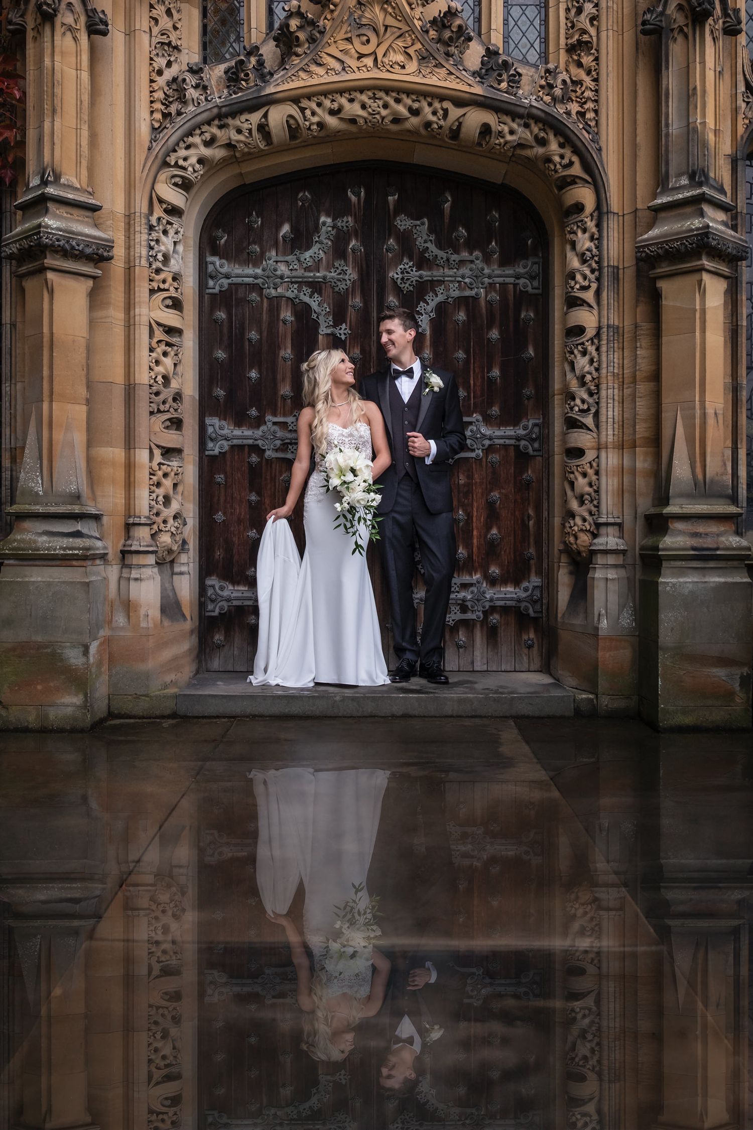 Wedding couple poses in front of ornate gothic doorway with reflection in puddle at historic building.