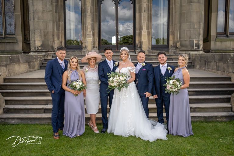 Large wedding party gathered for formal photos on lawn of stately stone building.