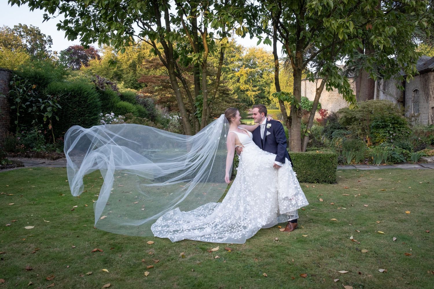 Wedding couple posing in a lush garden setting with flowing veil creating dramatic movement.