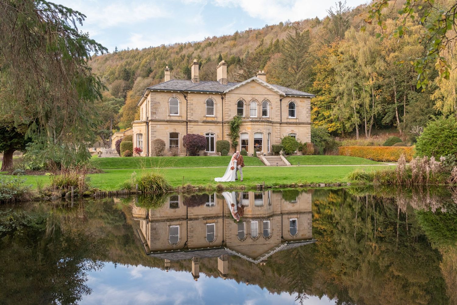 Panoramic view of a grand stone mansion reflected in a serene lake on an autumn day.