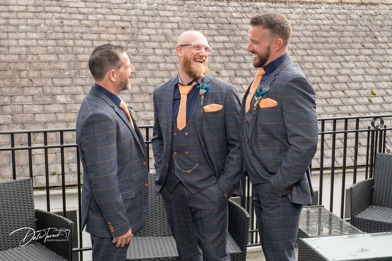 Three groomsmen in matching navy suits with orange pocket squares laughing together outdoors at a wedding venue.
