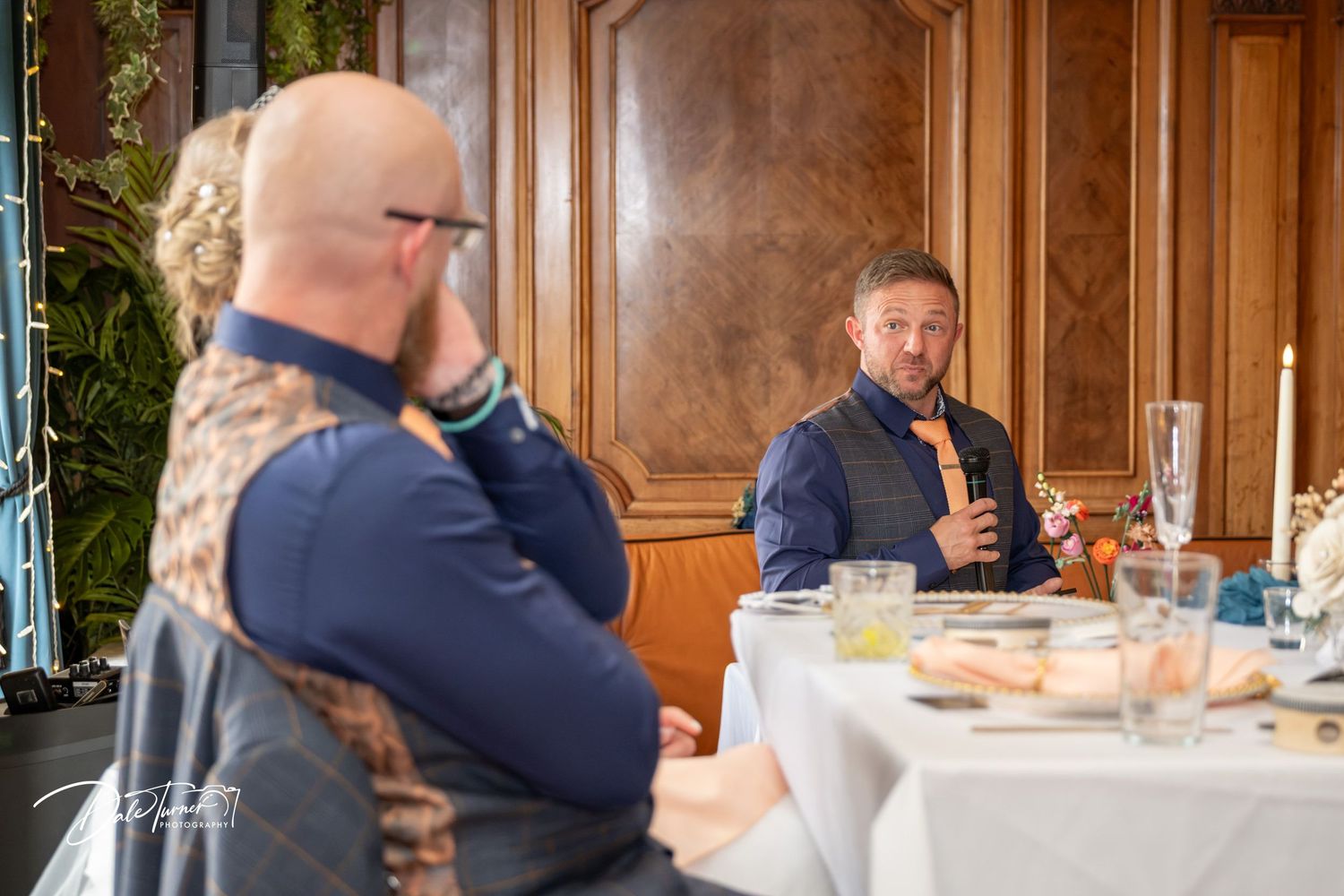 Two men in smart casual attire engaged in conversation at an elegantly set dining table in a wood-paneled room.
