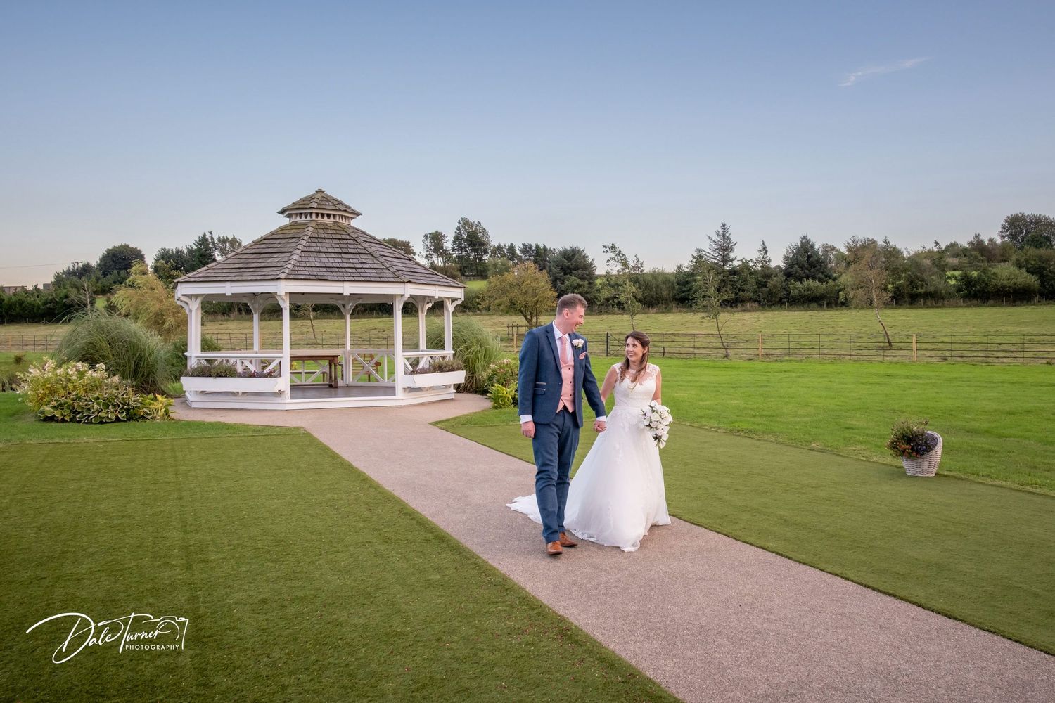 Panoramic view of wedding couple near white gazebo in an expansive garden setting.