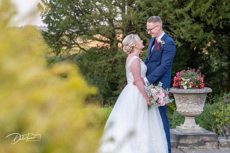 Elegant wedding couple poses by ornate garden urn filled with pink flowers.