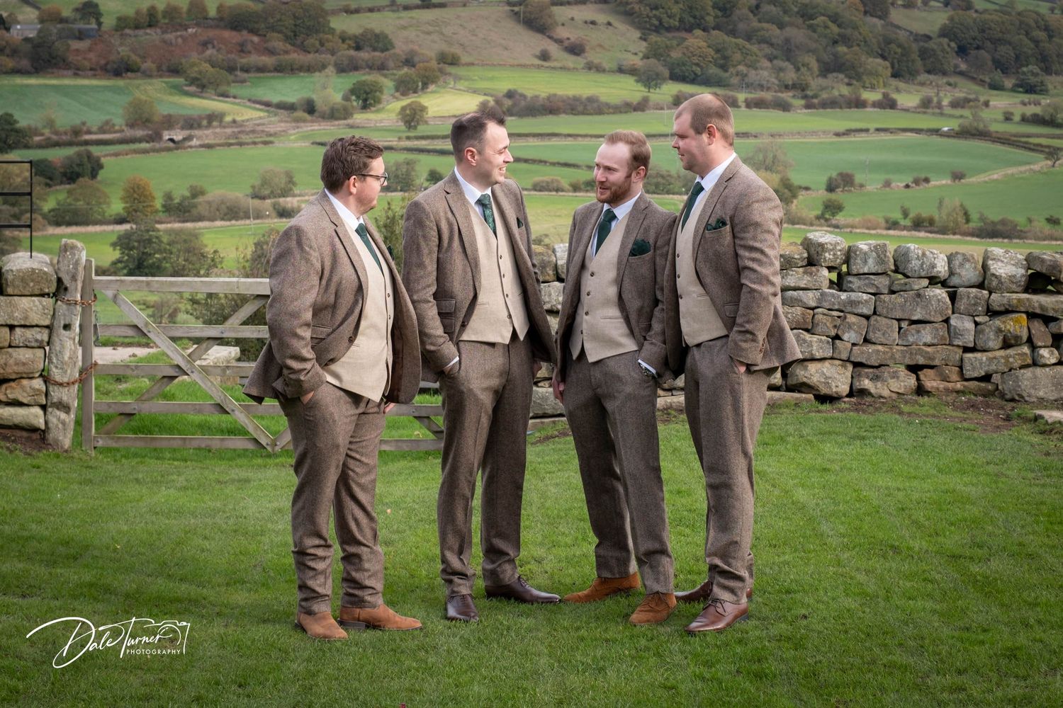Groomsmen in brown suits stand together on green grass with stone walls and rolling countryside hills in the background.