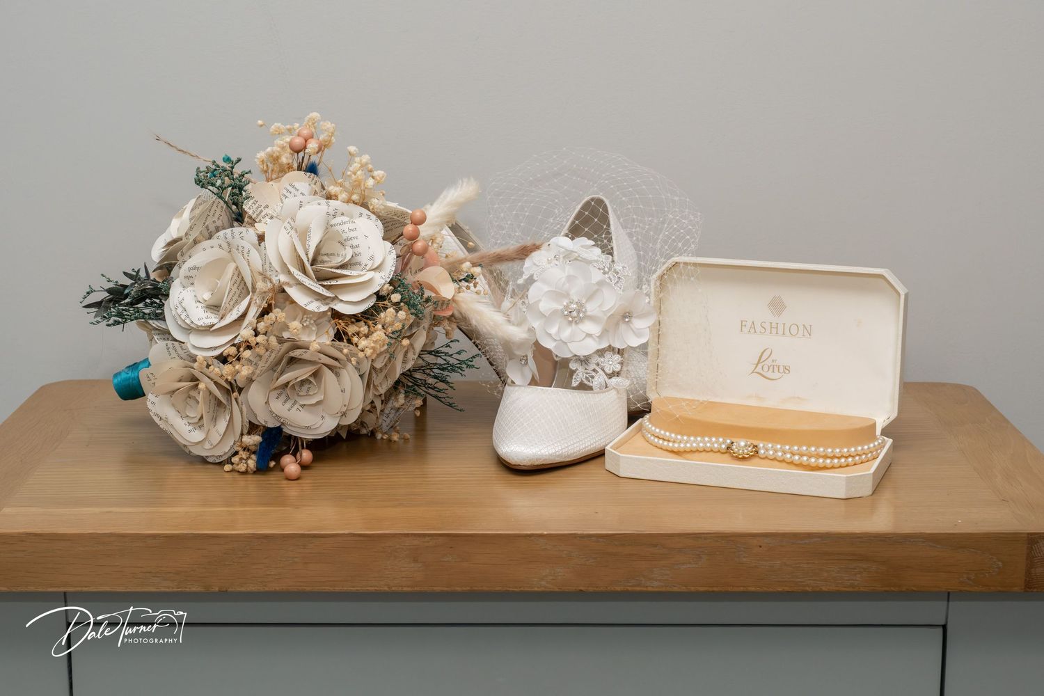 Wedding flat lay featuring a paper flower bouquet, white heeled shoe, pearl bracelet, and jewelry box on a wooden surface.