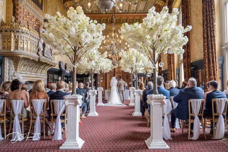 Grand hall wedding ceremony with ornate wood paneling and white cherry blossom decorations.