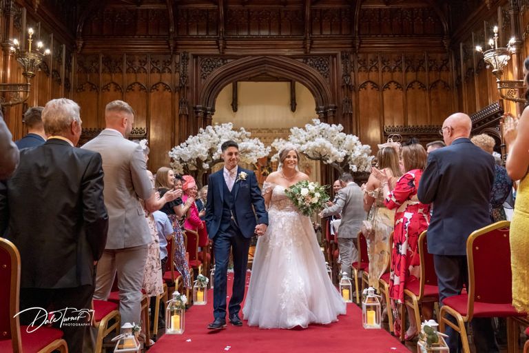 Indoor wedding ceremony in grand historic hall with ornate wooden details and red carpeting.