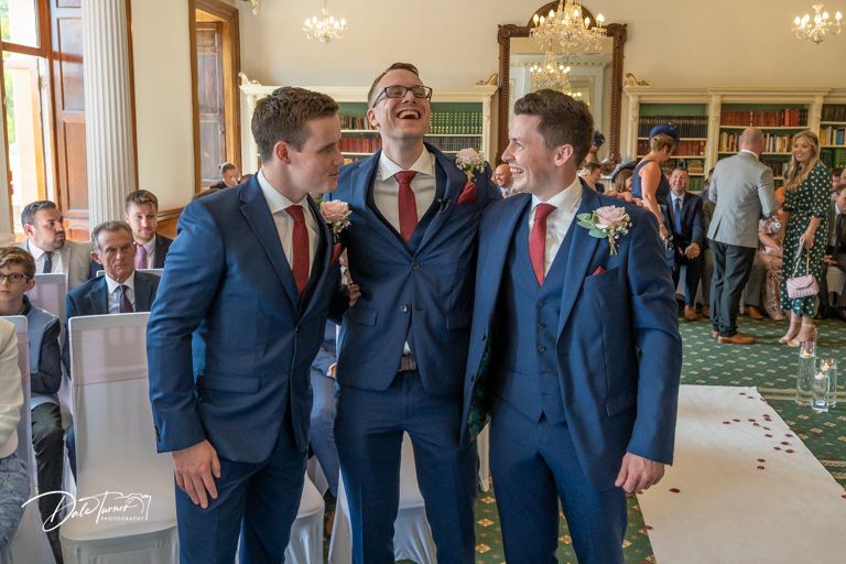 Groomsmen in navy suits gather during wedding ceremony in elegant indoor venue.