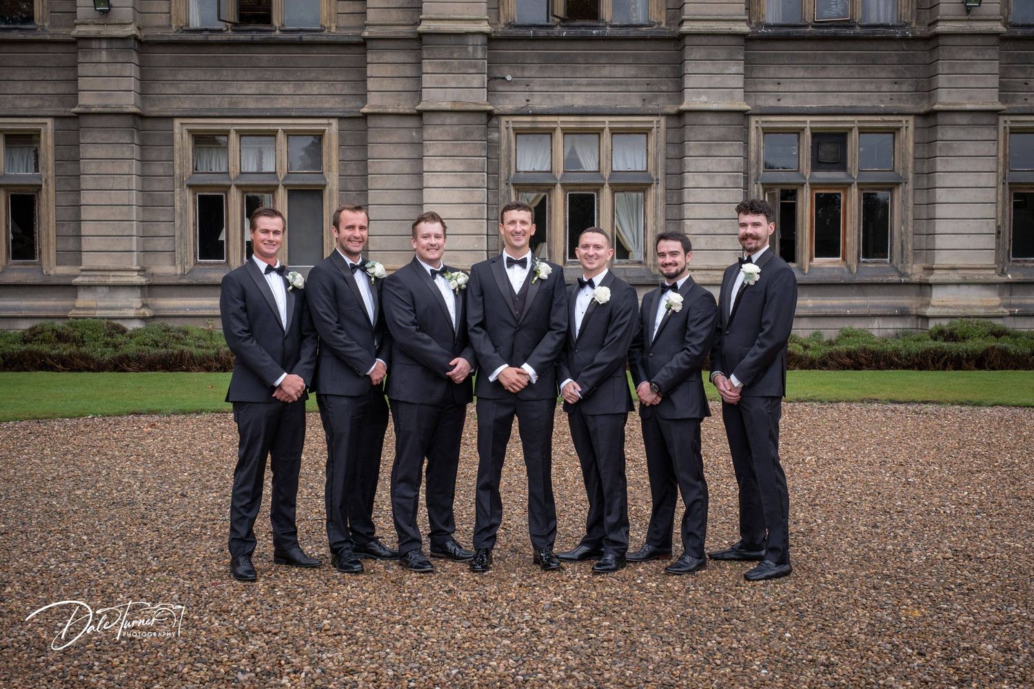 Groomsmen in formal black suits pose in front of historic stone building.