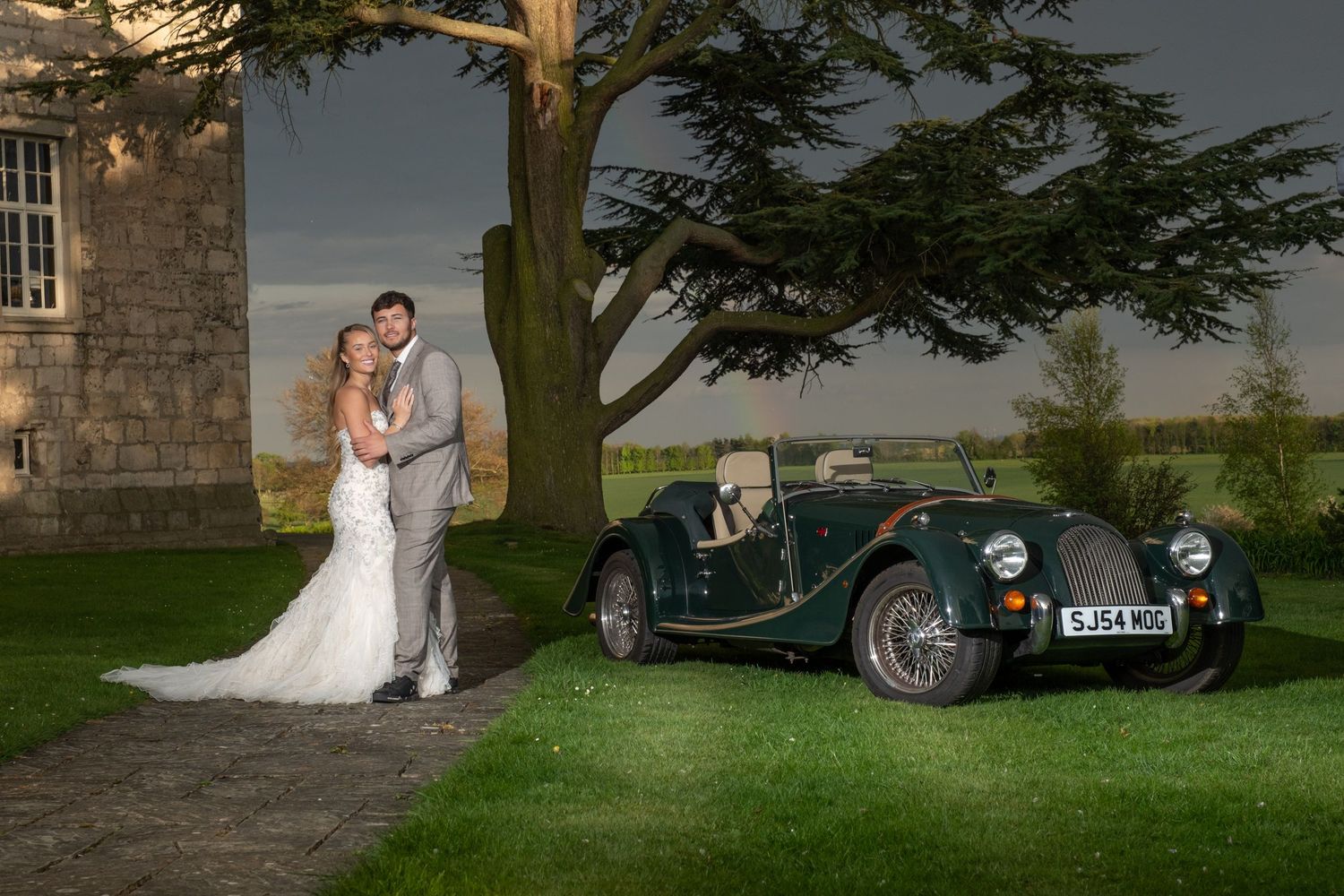 Elegant wedding photo shoot at dusk in front of a historic stone mansion with vintage cars.