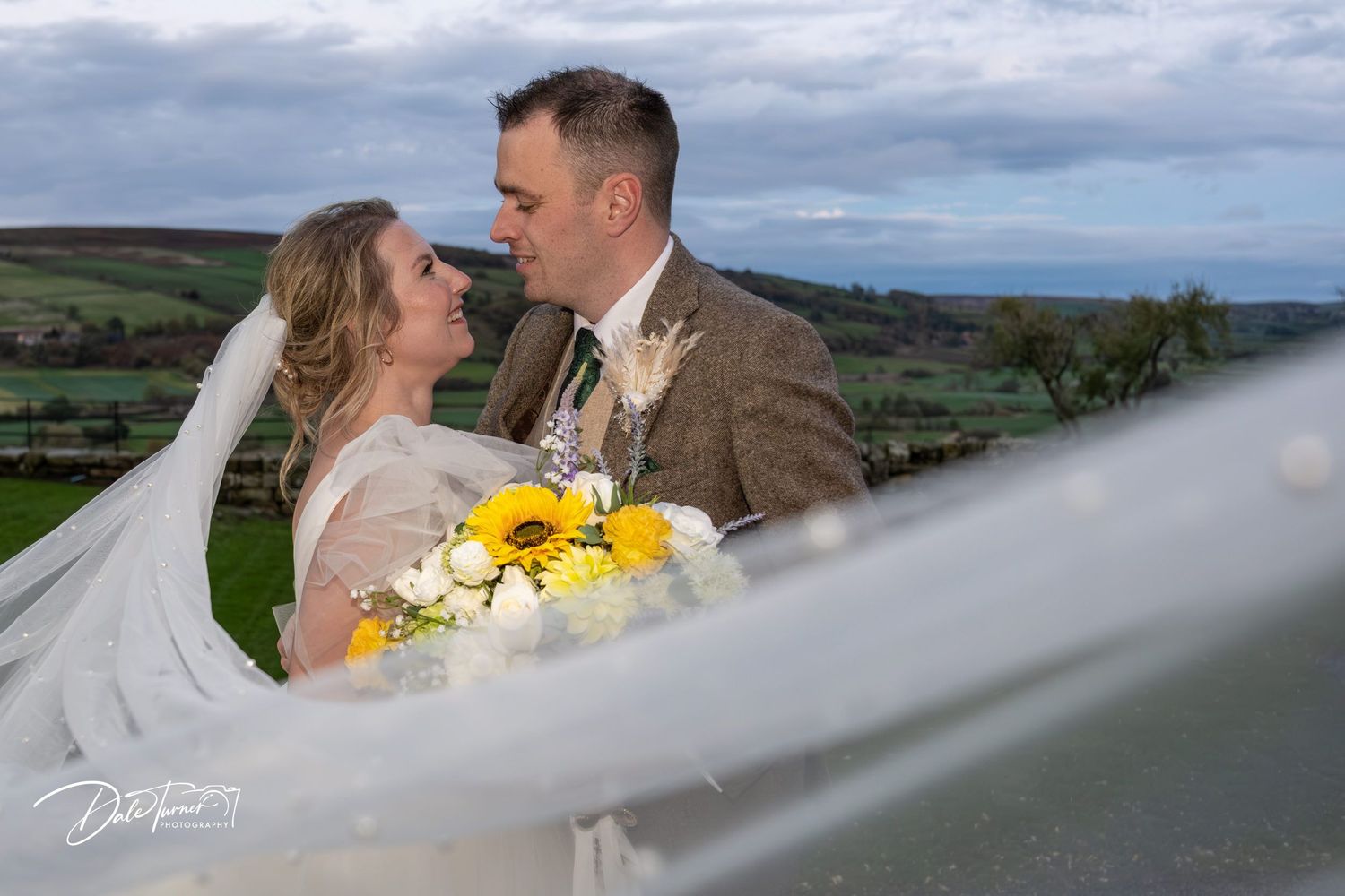 A romantic wedding photo with sunflowers against a scenic countryside backdrop with rolling hills and cloudy skies.