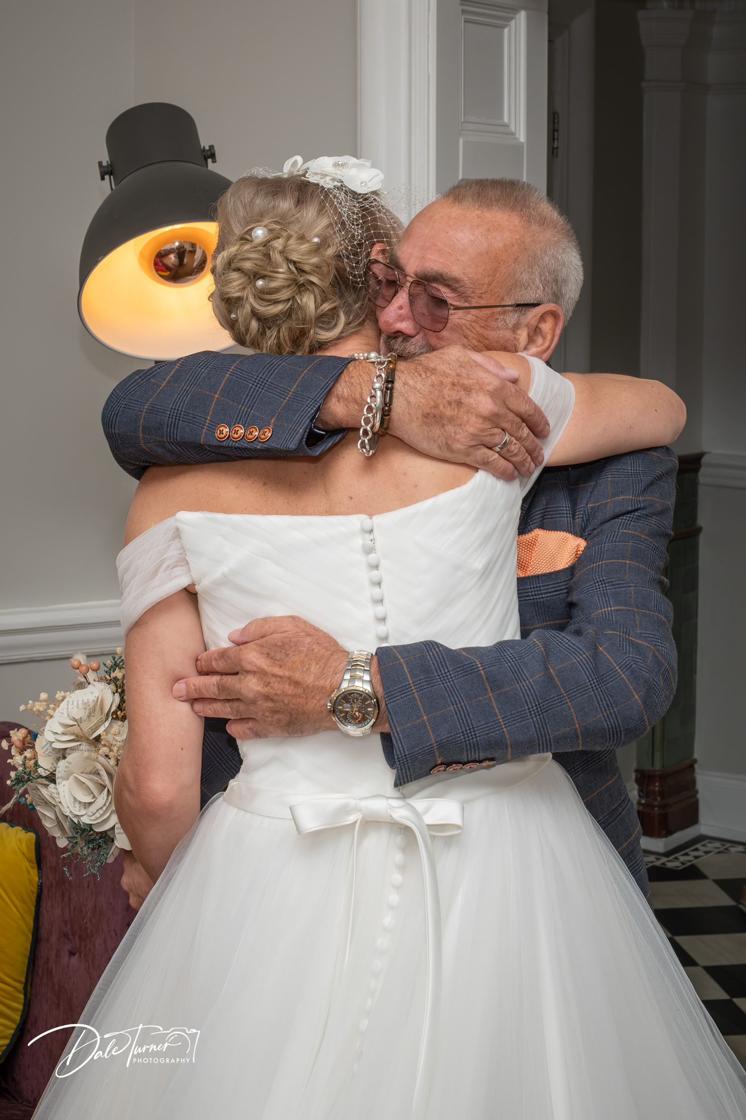 Elderly man in blue tweed suit embracing bride in white wedding dress during emotional wedding ceremony hug.