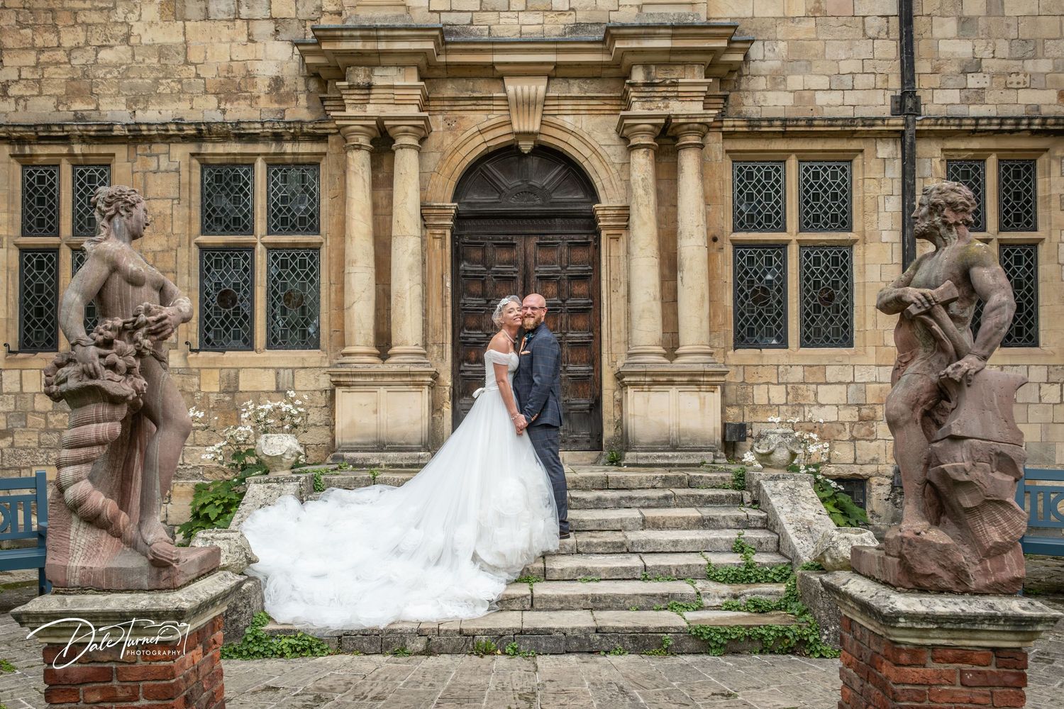 Bride and groom pose on stone steps flanked by classical statues outside a historic stone building.