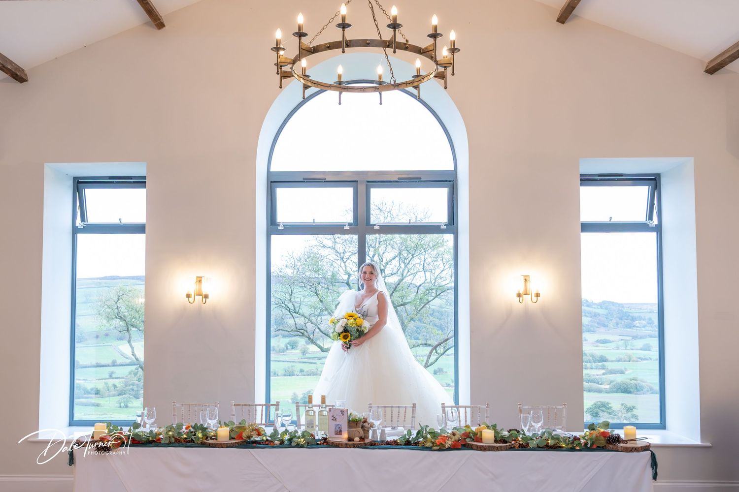 A bride stands silhouetted in front of an arched window with wall sconces and a rustic chandelier in an elegant venue.