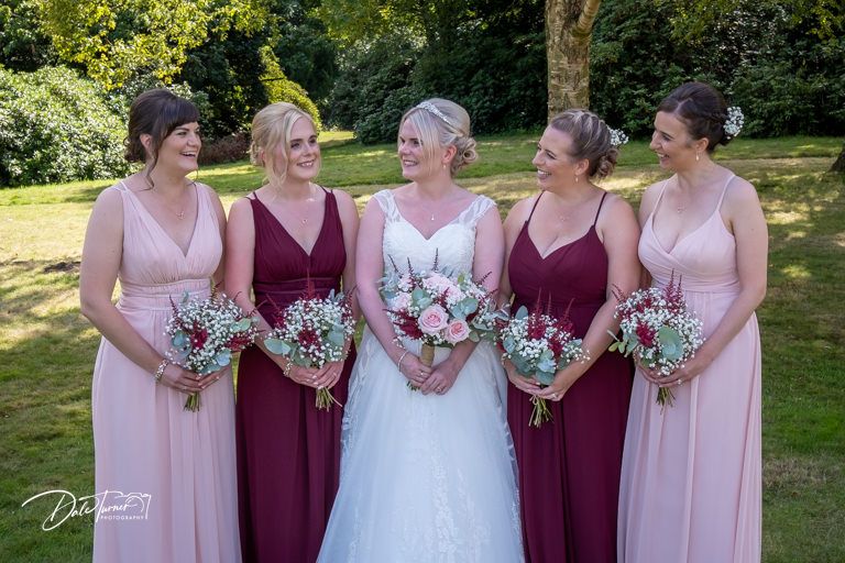 Bridesmaids in burgundy and pink dresses stand together holding bouquets in an outdoor garden setting.
