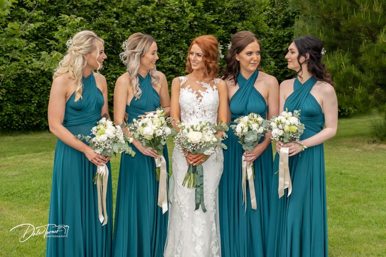 Bridesmaids in teal dresses pose with bouquets in garden setting.