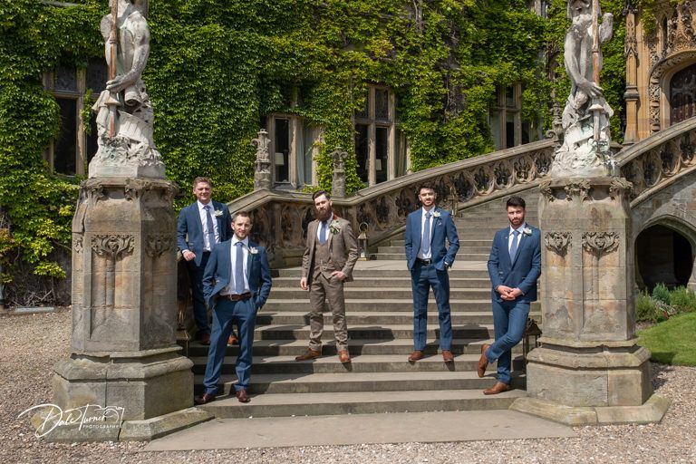 Wedding party poses on grand stone steps surrounded by lush greenery at historic venue.