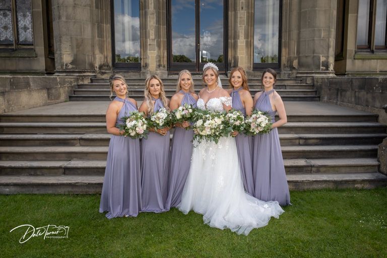 Wedding group stands outside historic stone building with classic architecture and manicured lawn.
