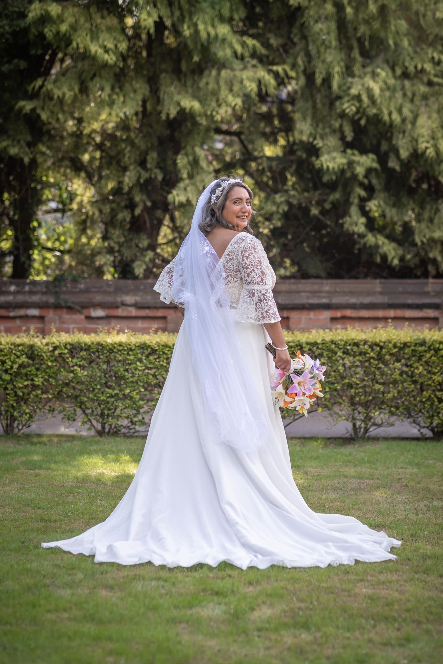Two brides in white wedding dresses pose together on manicured garden grounds.