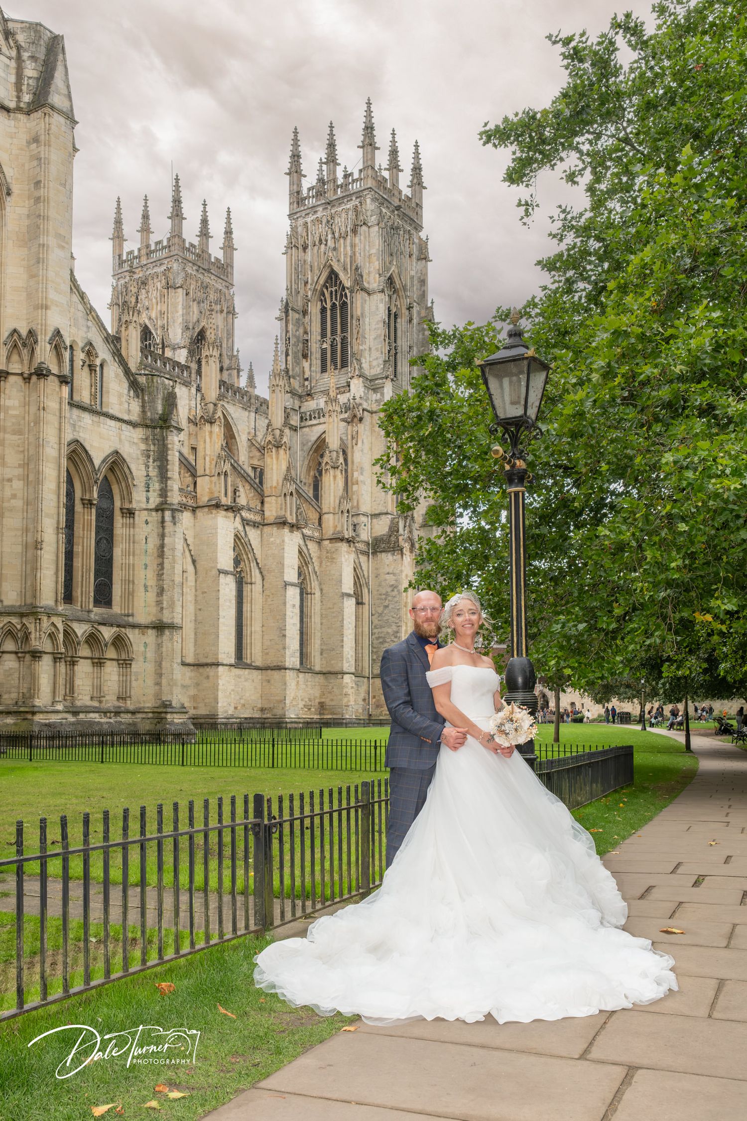 Bride and groom posing in front of York Minster Cathedral on their wedding day, with lush green trees nearby.