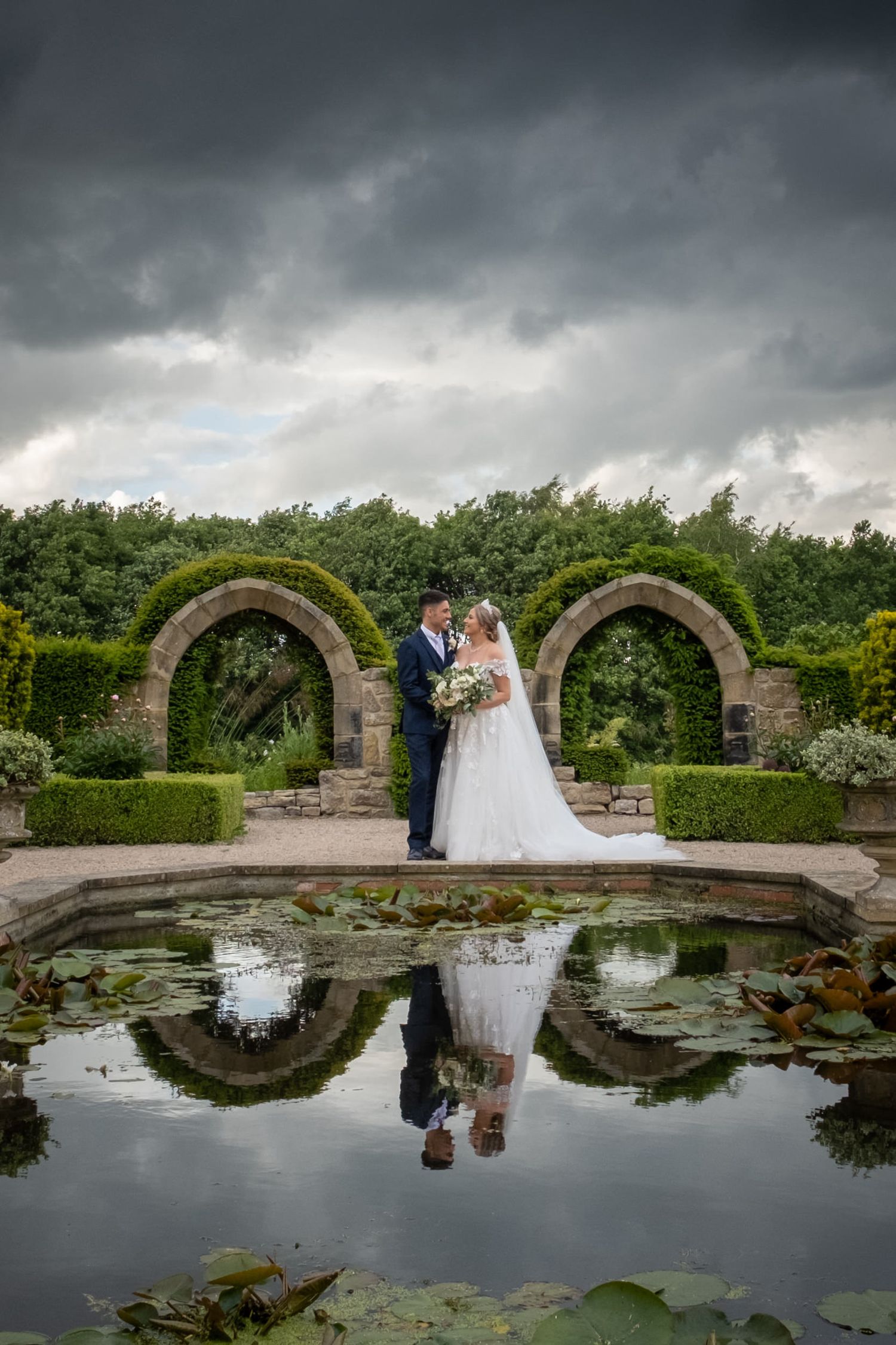 Wedding couple by stone arches reflecting in lily pond under cloudy sky.