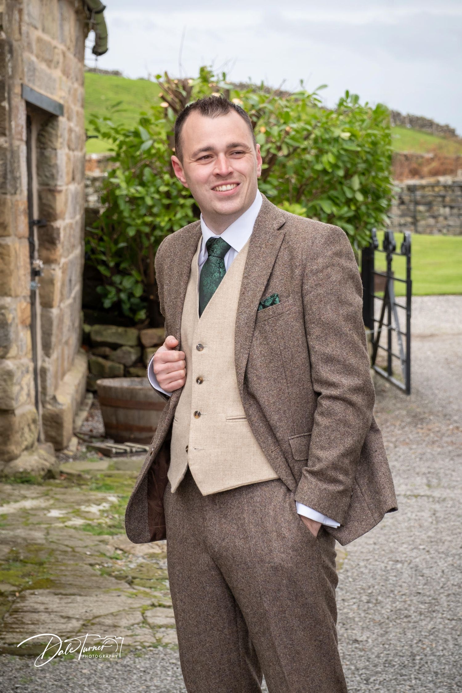 A groomsman in a brown tweed suit and beige vest poses against stone archways at a rustic outdoor venue.