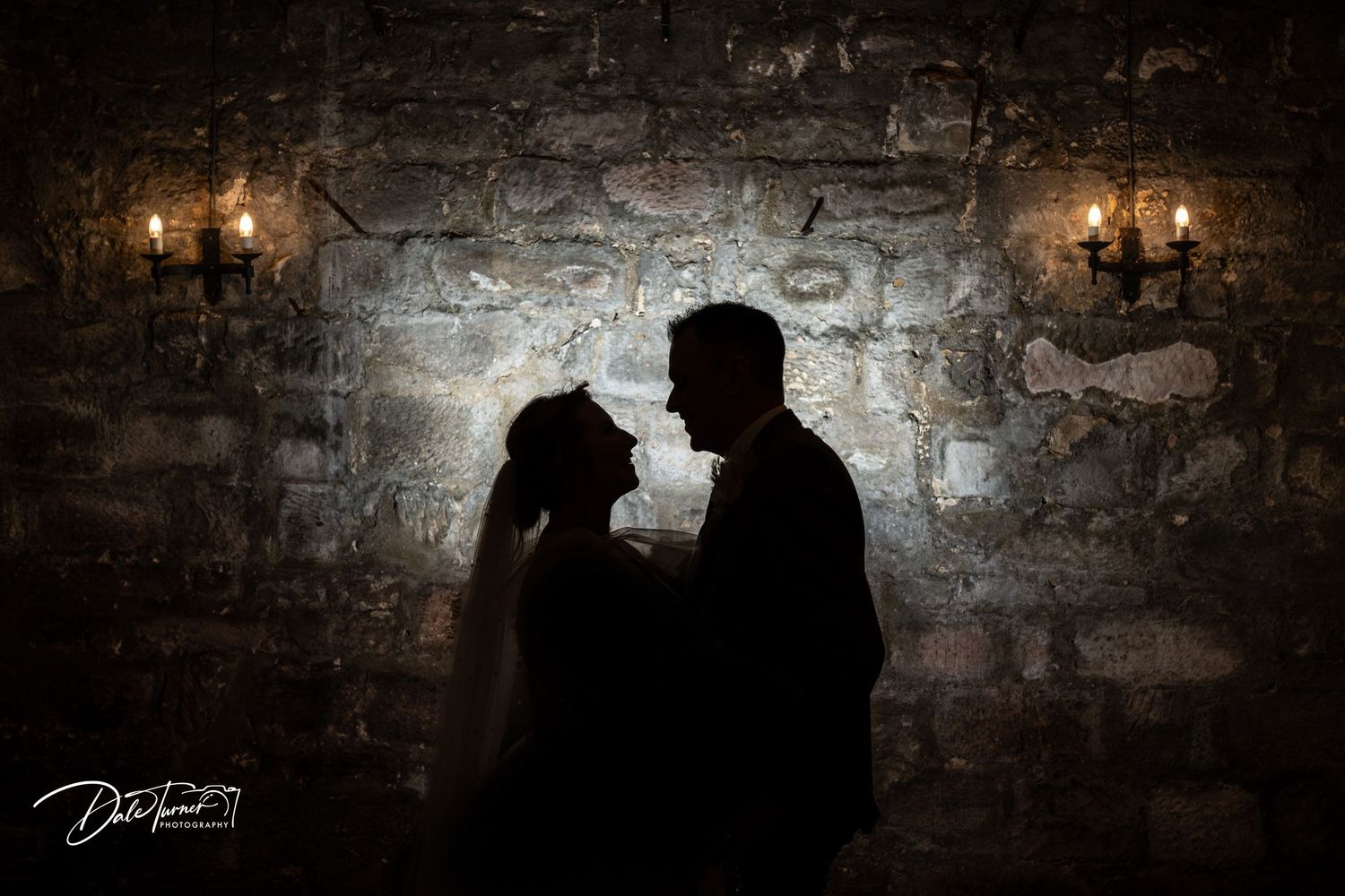 Silhouetted couple stands intimately against rustic stone wall with glowing vintage sconces creating romantic ambiance.