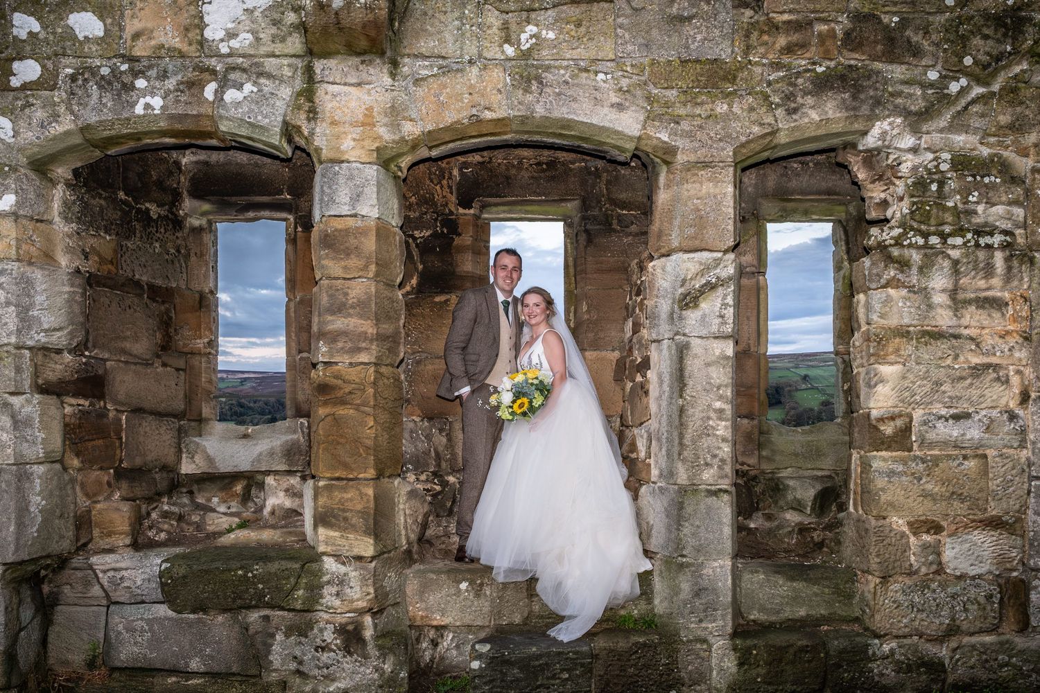 A series of stone archways frame scenic views of a wedding dress against a rustic stone wall backdrop.