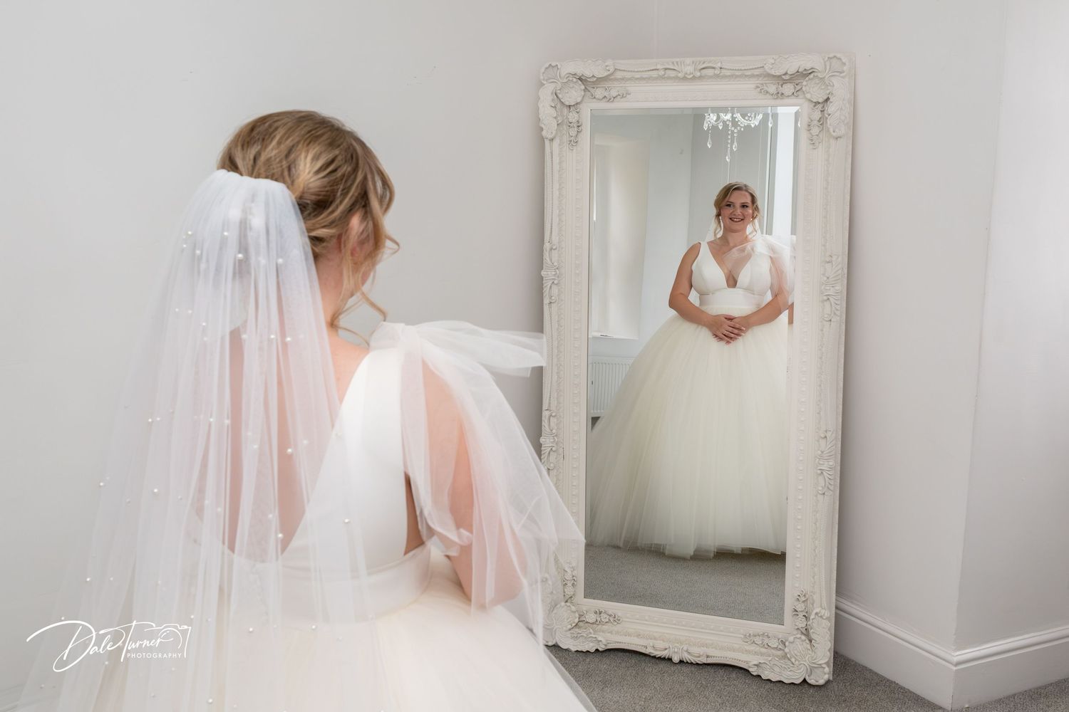 A bride in a flowing white gown and pink veil poses elegantly in a light-filled room with ornate decor.