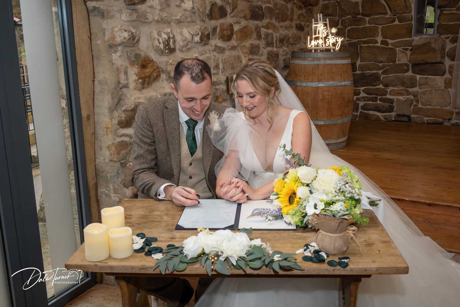 A group of people signing documents at a rustic wooden table with stone walls and sunflowers in the background.