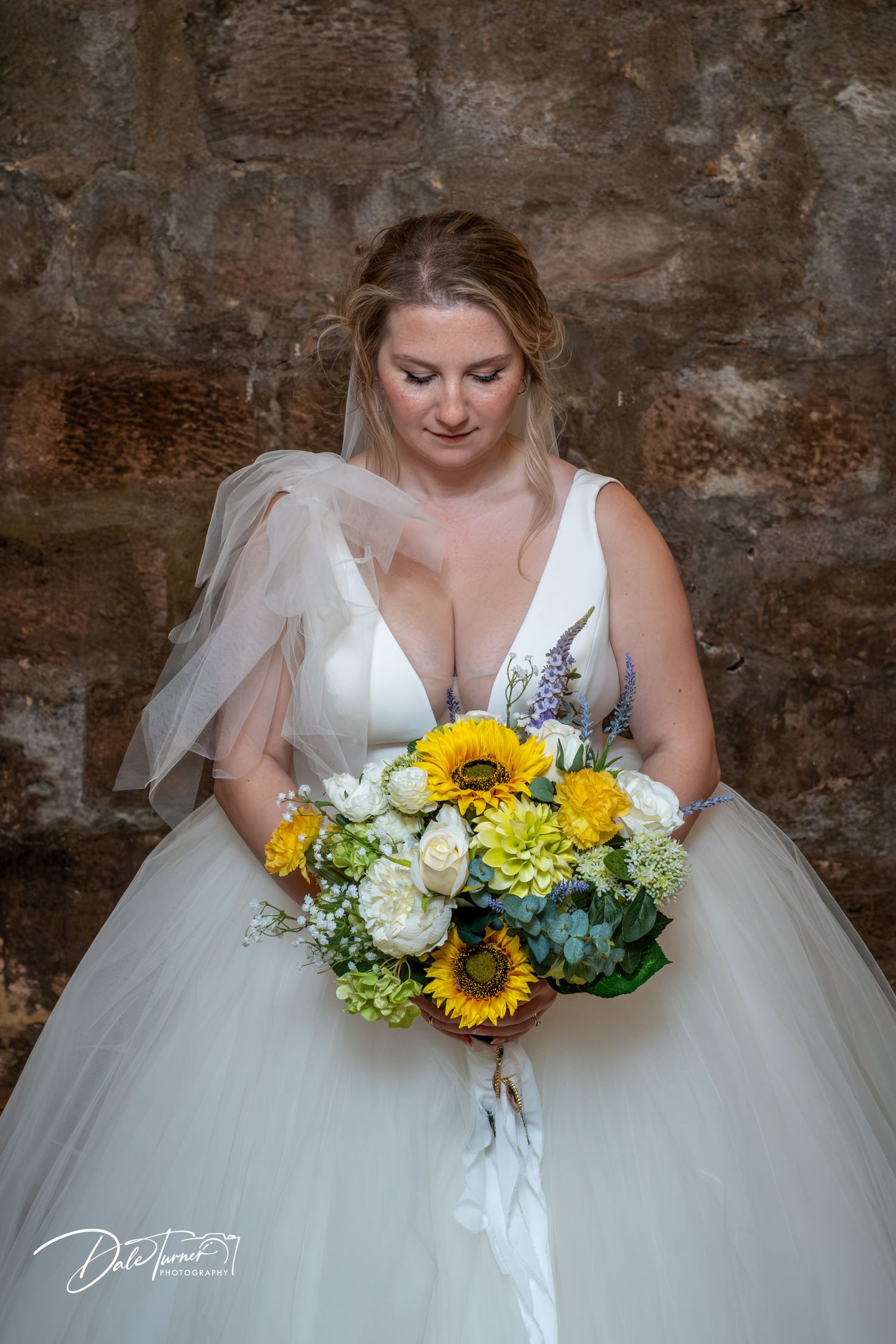 A bride in a white wedding gown holds a vibrant bouquet with sunflowers against a rustic brick wall background.
