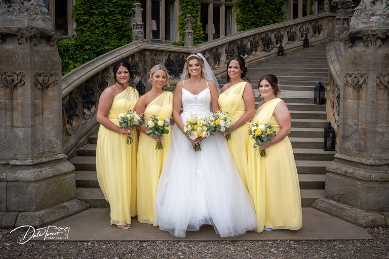 Bridal party in yellow and white dresses standing together on ornate stone staircase.