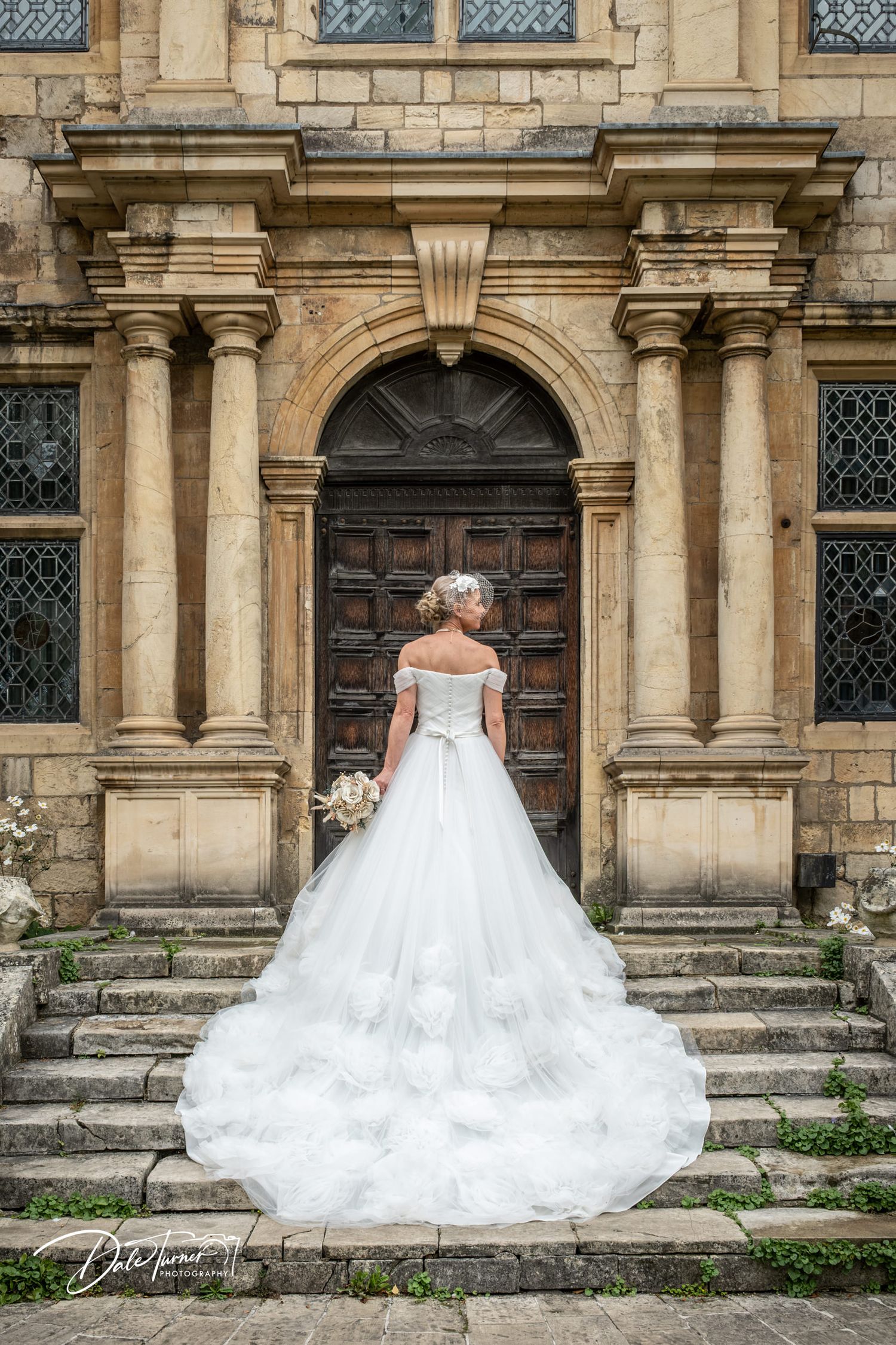 Bride in elegant white ballgown with long train stands on stone steps before ornate arched doorway of historic manor.