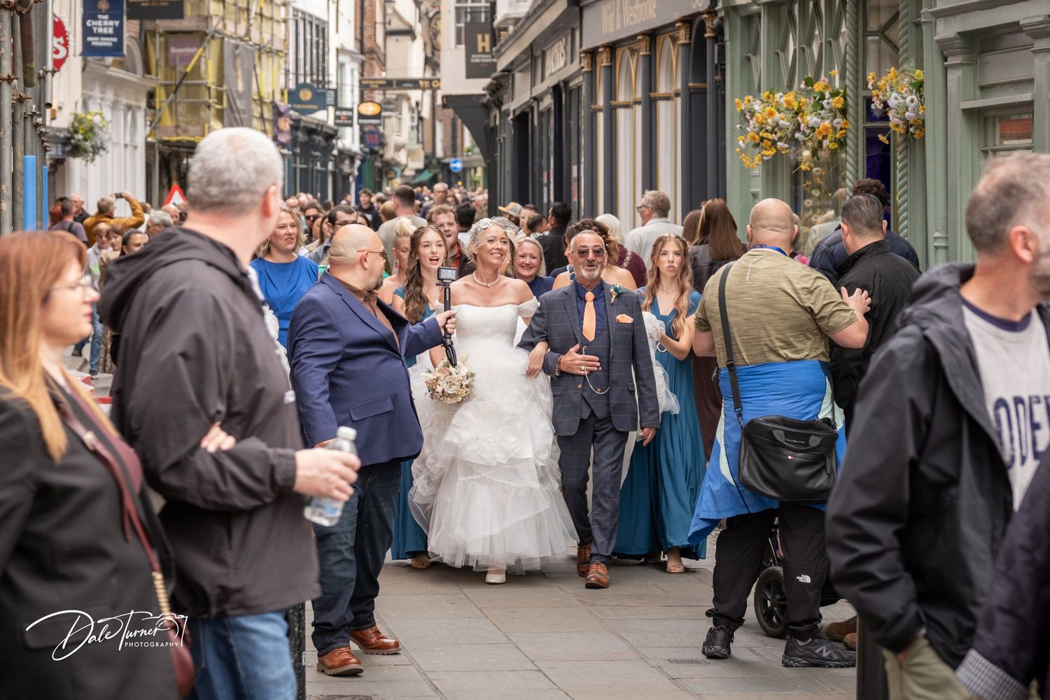 Bride in white gown walks through busy pedestrian street with wedding guests during outdoor celebration.