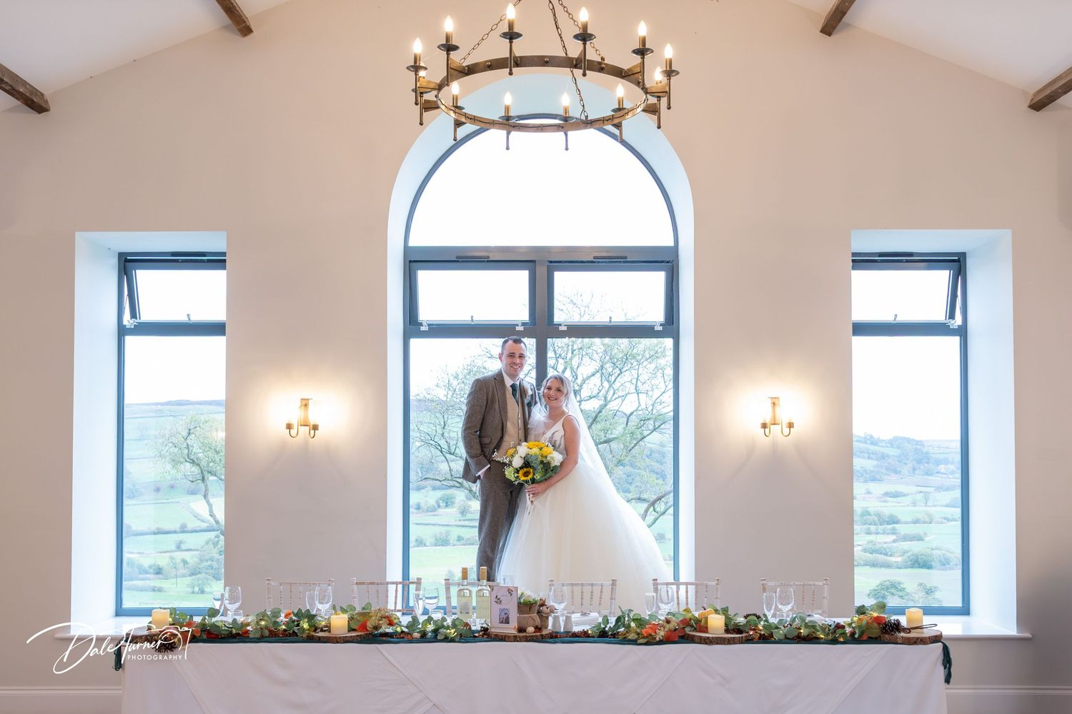 Elegant rustic wedding venue interior with exposed beams, chandeliers, and arched windows decorated with floral arrangements.