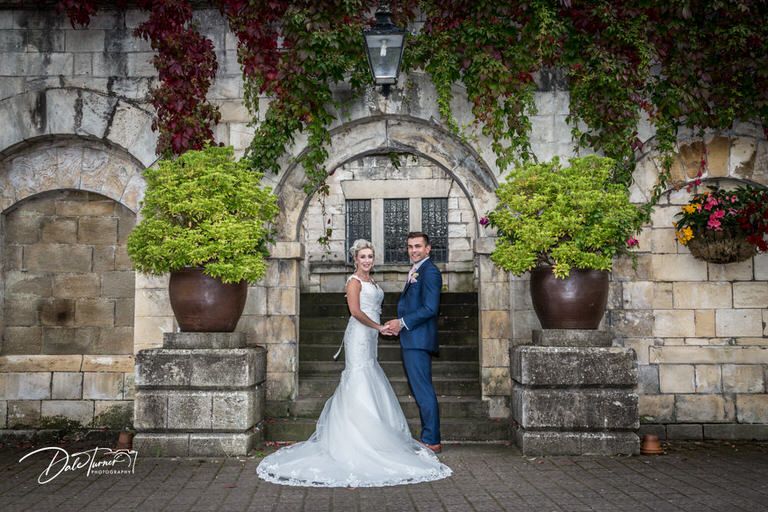 Elegant wedding couple poses against historic stone building with climbing vines.