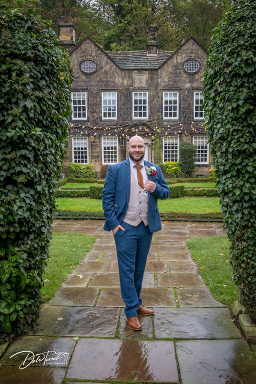 Wedding party member poses in a navy suit against historic stone building with ivy and windows.