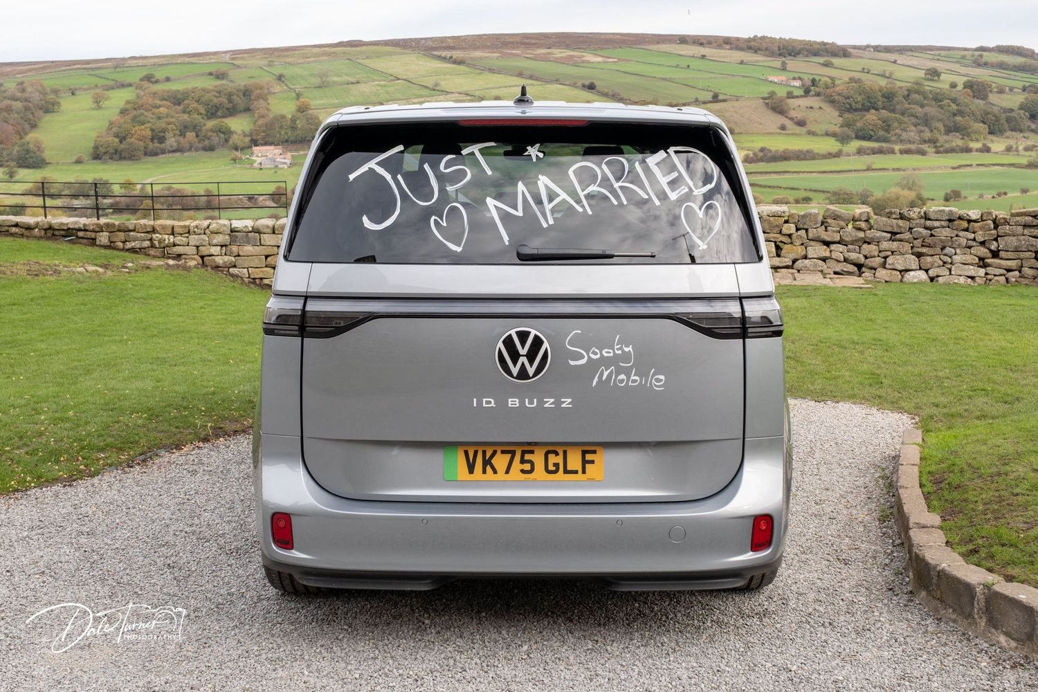 A grey Volkswagen van with 'Just Married' written on the back window parked in front of rolling green hills.