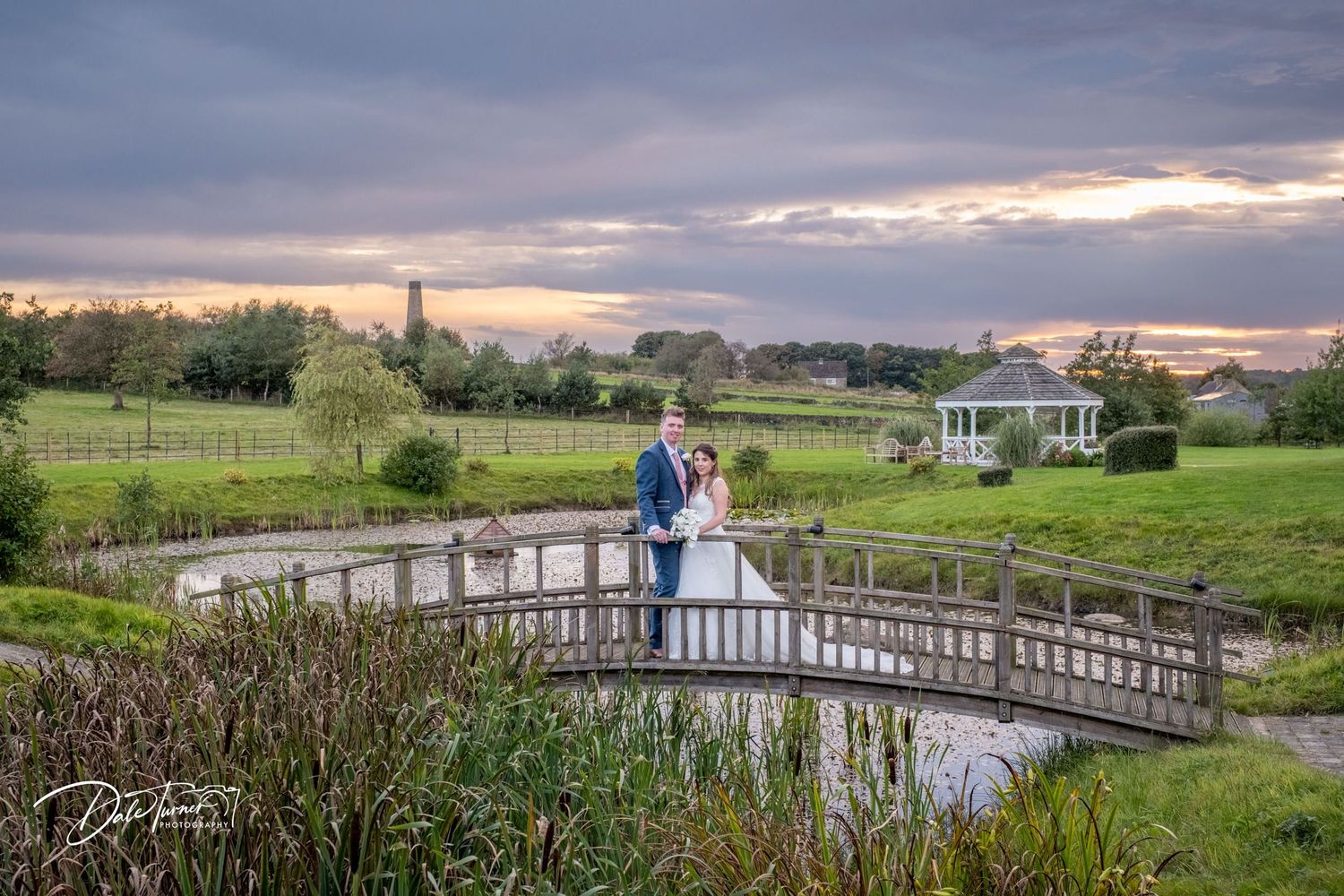 Romantic sunset wedding photos captured on wooden bridge over tranquil pond with gazebo in background.