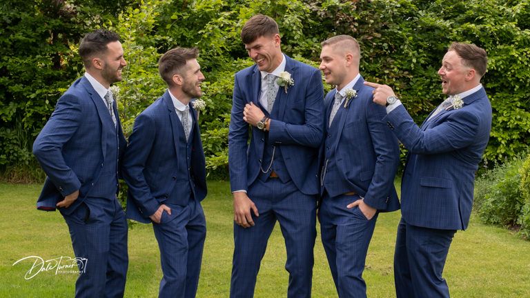 Groomsmen in navy suits share laughs and casual moments on a sunny lawn.