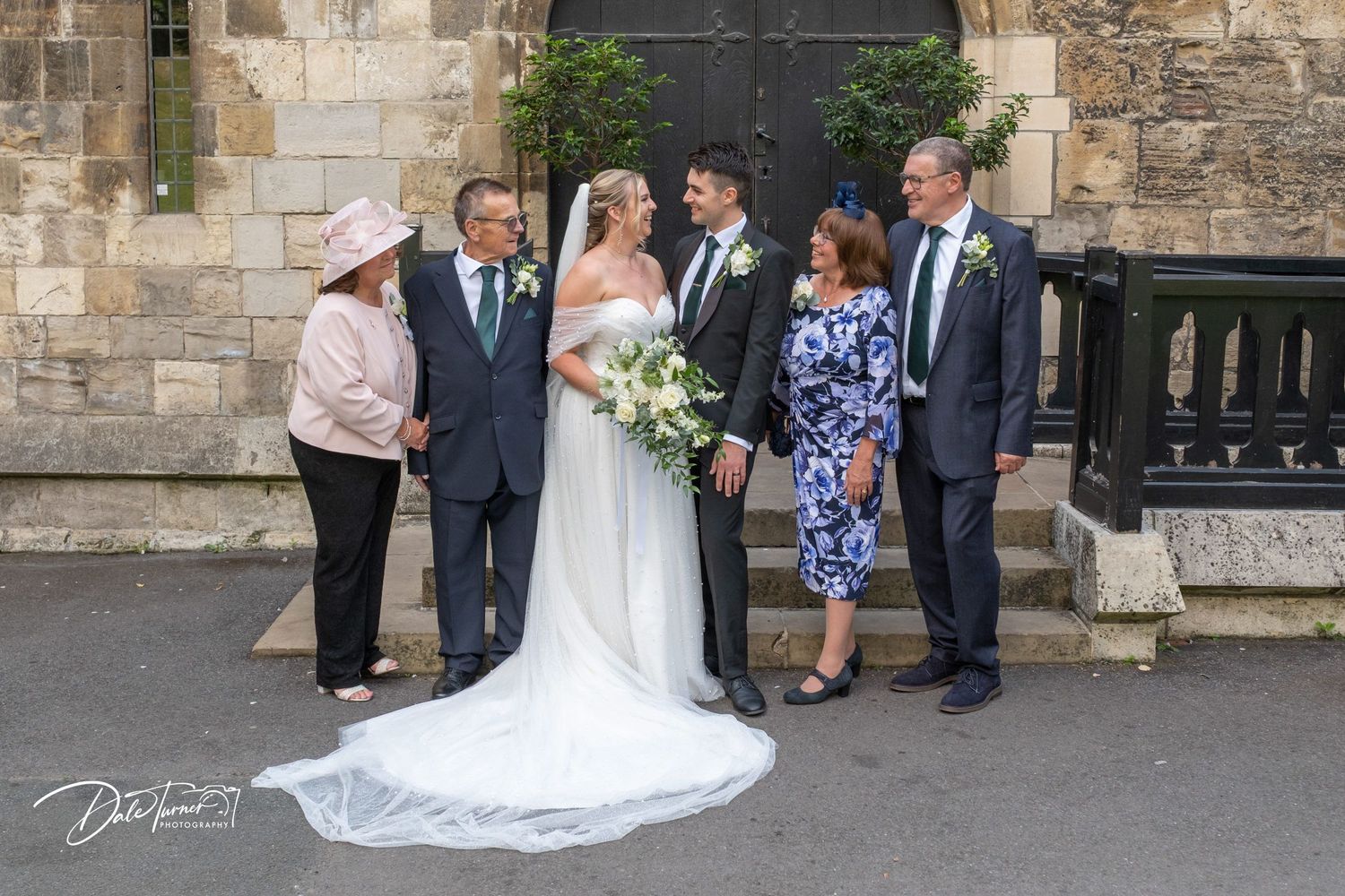 Wedding party poses outside historic stone church with bridesmaids in emerald green dresses and white floral bouquets.