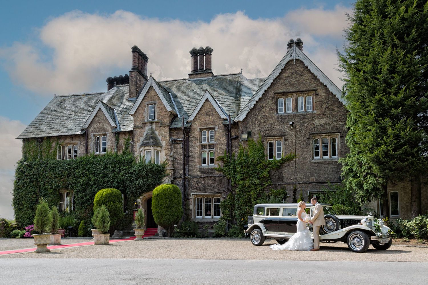 Gothic style stone manor house exterior with vintage cars parked in front.