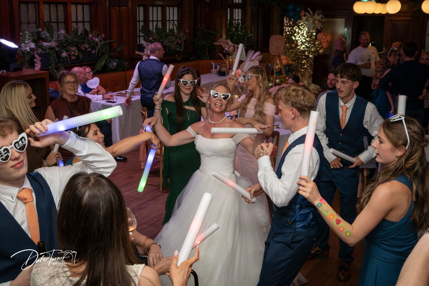 Bride and groom dancing with guests holding glow sticks at a lively wedding reception party.