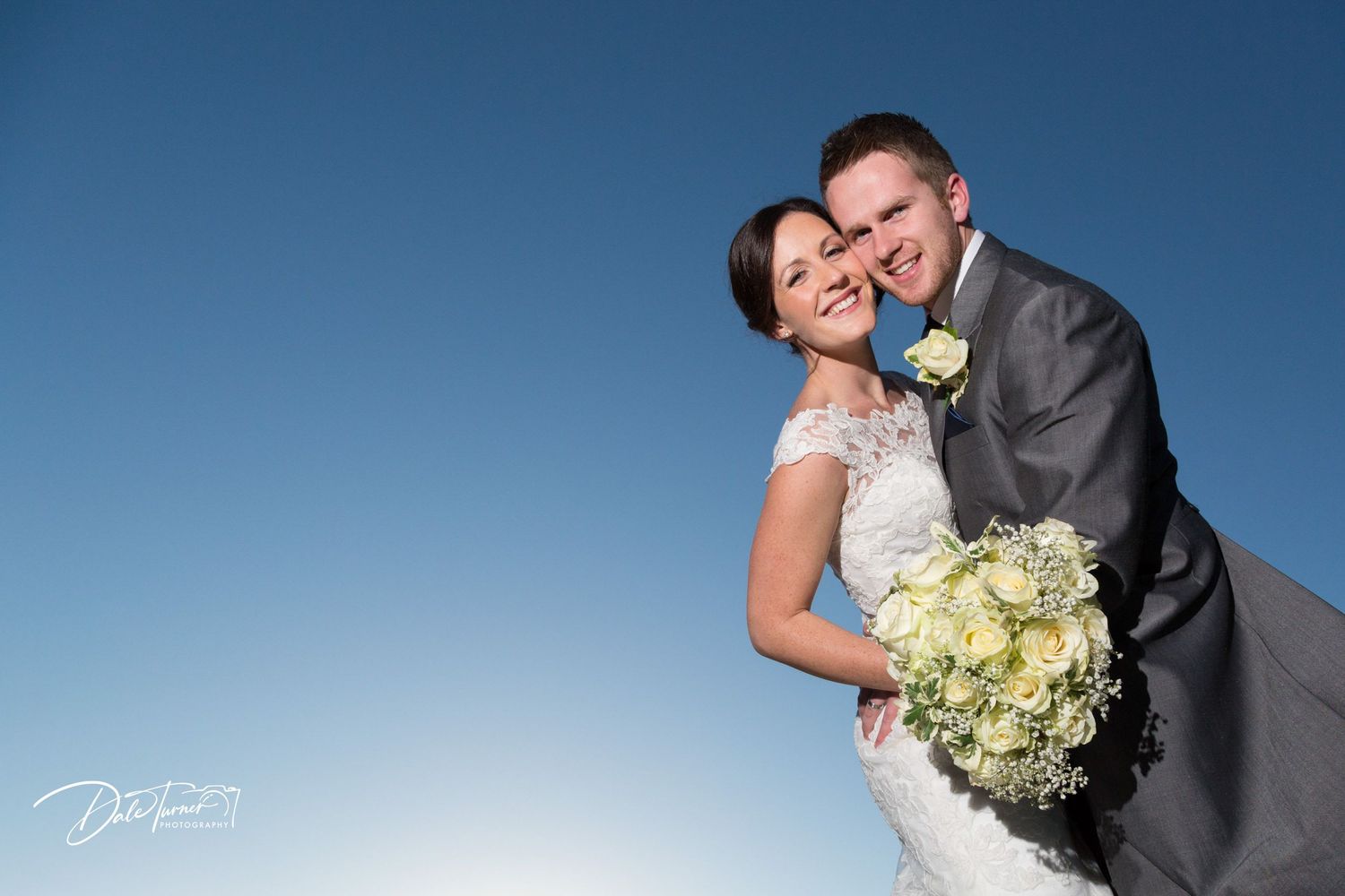 Romantic wedding portraits against bright blue sky with white floral bouquet.