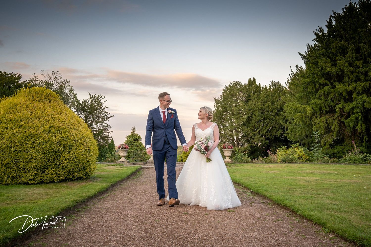 Wedding couple walks along a scenic countryside path during sunset.