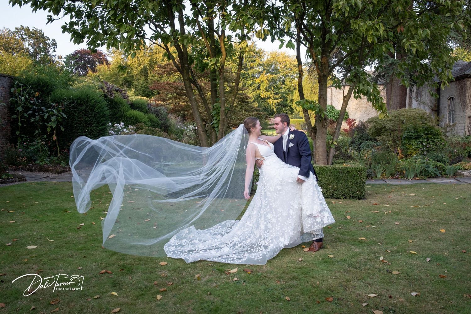 Elegant wedding couple poses in a garden setting with flowing white veil and autumn foliage.