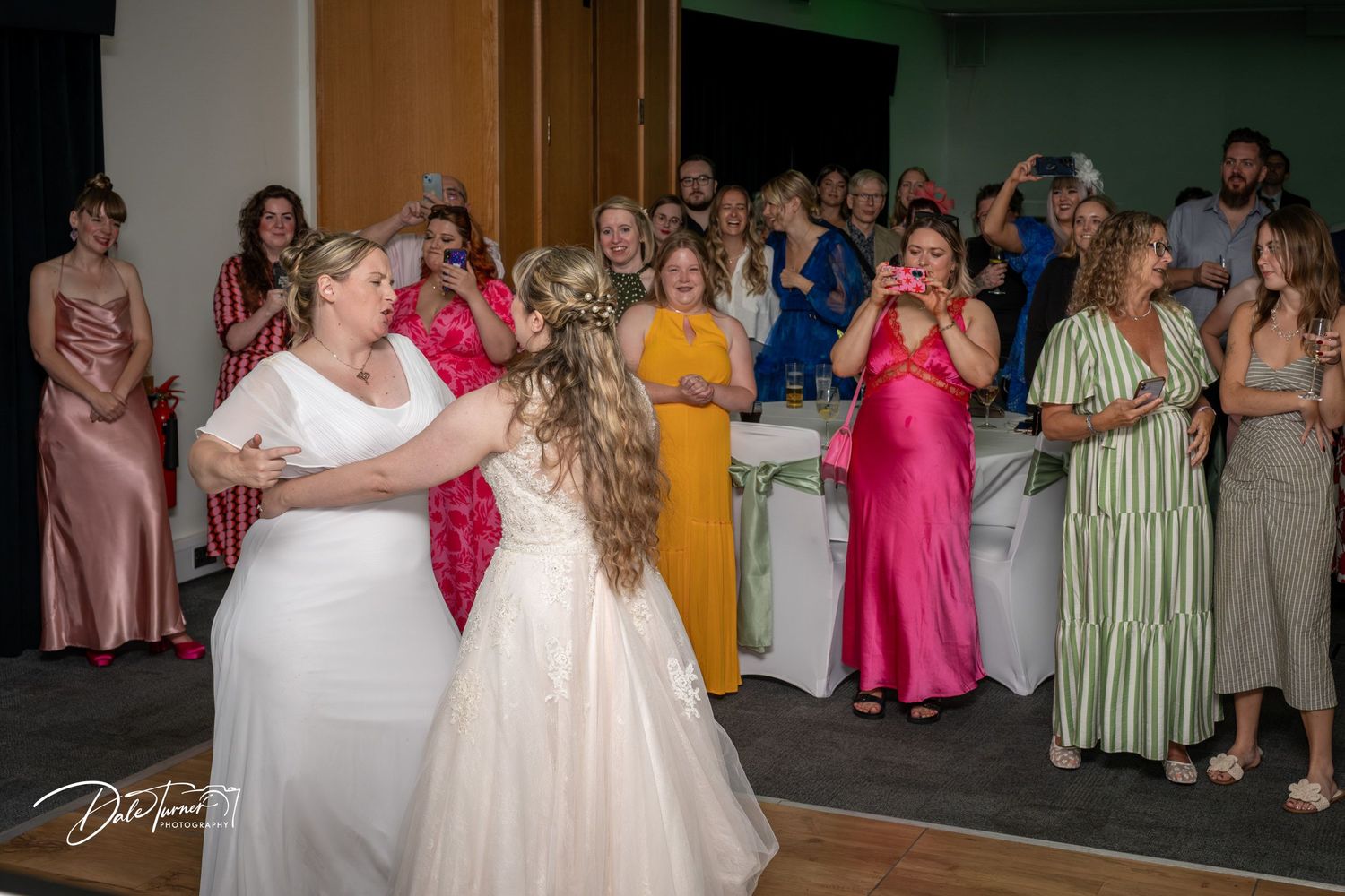 Wedding couple dancing together surrounded by guests in bright formal dresses at a reception venue.
