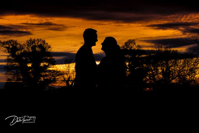 A silhouetted couple against a dramatic orange sunset sky with trees in the background.