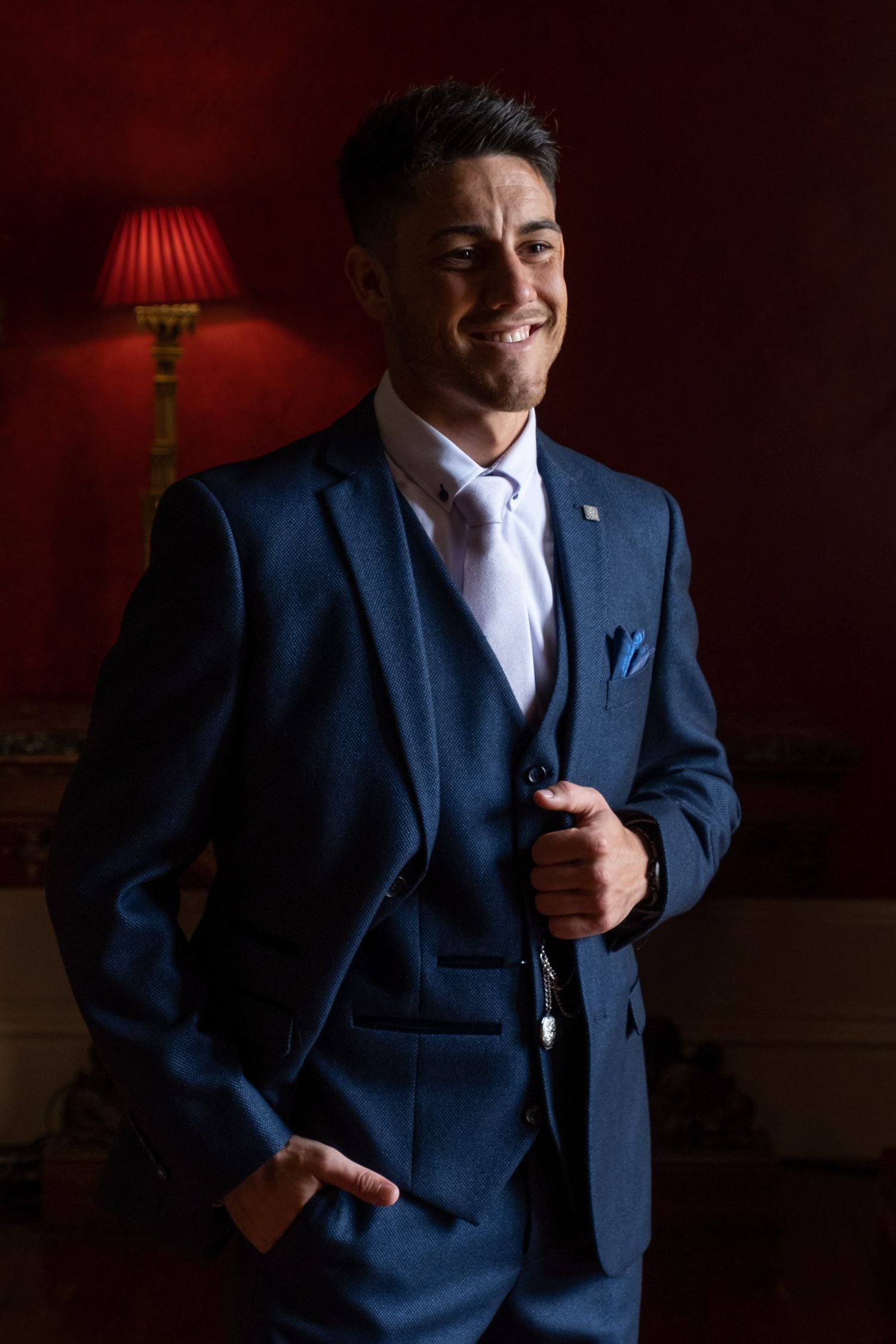 Dramatic portrait of groom in navy suit against deep red wall with lamp lighting.